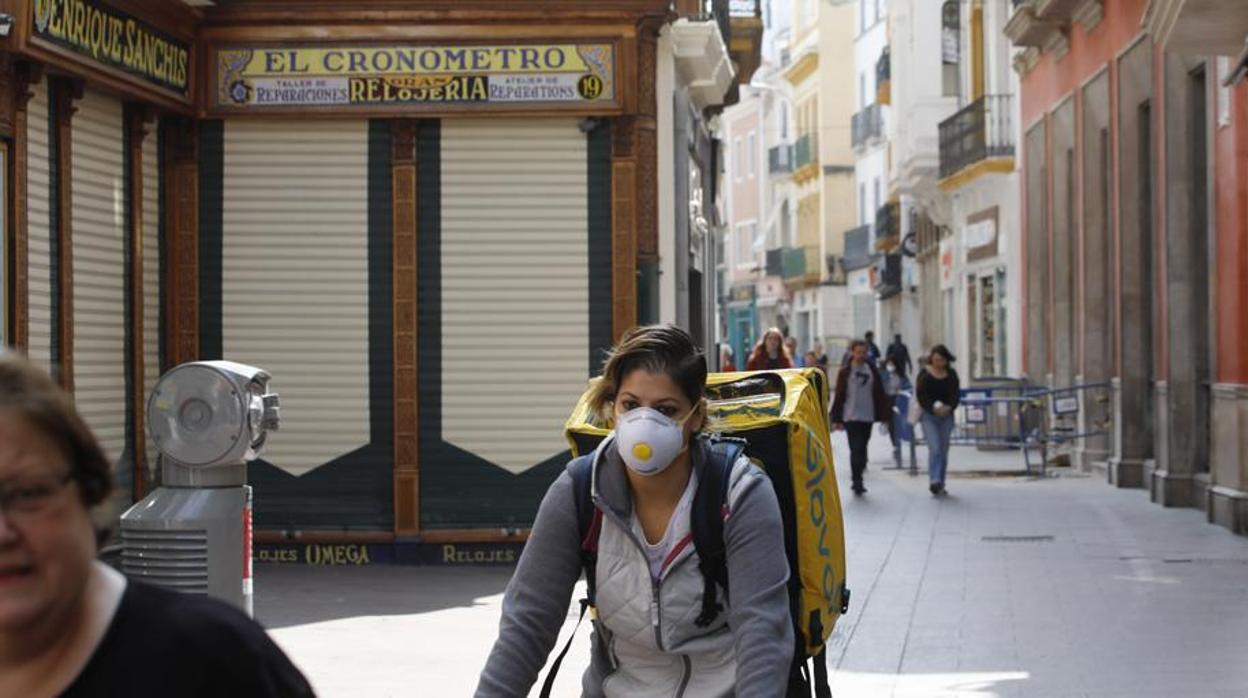 Una trabajadora de Glovo pasa por una calle del centro de Sevilla con los comerccios cerrados