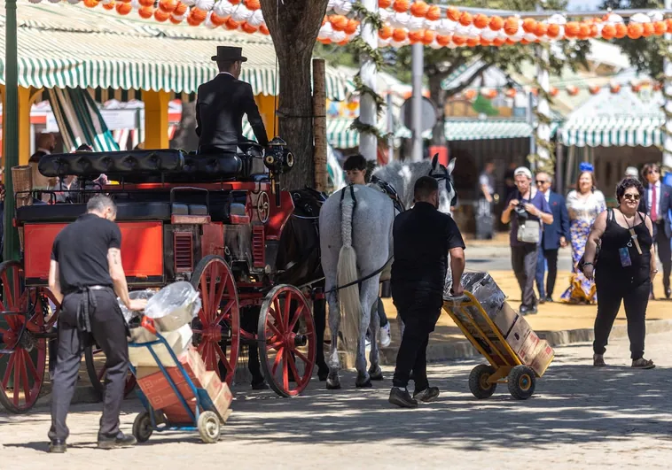 Dos camareros cruzan la Feria de Abril con material de hostelería, junto a coche de caballos