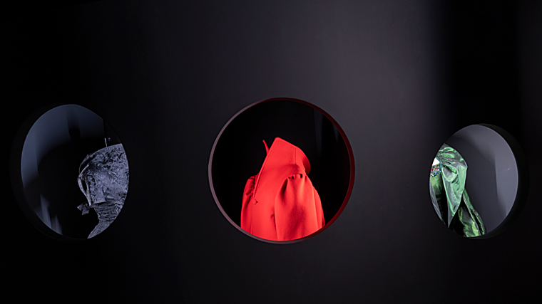 Vitrina con ventanas de ojo de buey para ver en con detalle los cuellos de las piezas. De izq. a dcha.: capa de terciopelo de seda morado de tejido drapeado con cuello grande en volante, costuras que conectan con la espalda y mangas japonesas con puños, modelo 145 de la colección de invierno de 1951, de Cristóbal Balenciaga. Abrigo rojo de fieltro con escote caído en la espalda, cuello alto forrado para resaltar el rostro y mangas 'oversize' fruncidas, es el modelo 65 de la colección de invierno 2019, firmado por Demna para Balenciaga. Conjunto de noche verde escarabajo iridiscente compuesto por una capa con solapa ancha y fruncida que rodea el cuello y vestido con cuello barco y mangas tres cuartos, bordadas con cuentas doradas, 'strass' rosa y cristales esmeralda, modelo 102 de invierno de 1961, de Cristóbal Balenciaga