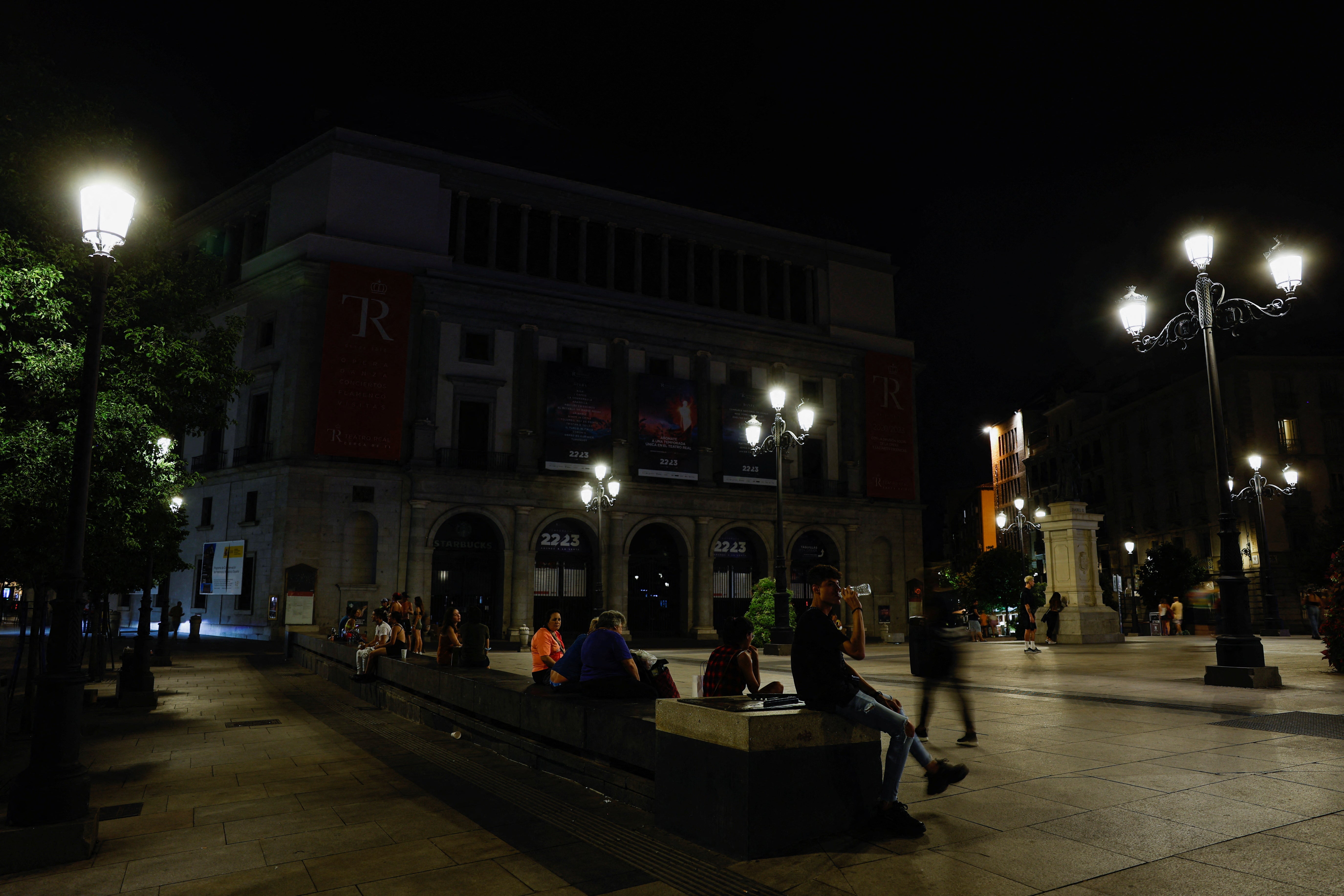 Un grupo de personas descansan en Ópera, con el Teatro Real con las luces apagadas.