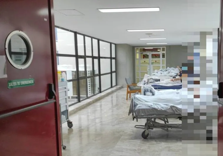Camas con pacientes en un pasillo sin cortinas entre Urgencias y Consultas Externas en el hospital de la Ribera