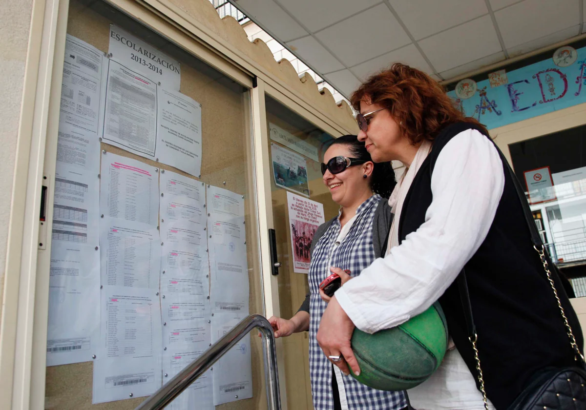 Dos mujeres consultan un tablón de un colegio con listados de escolarización