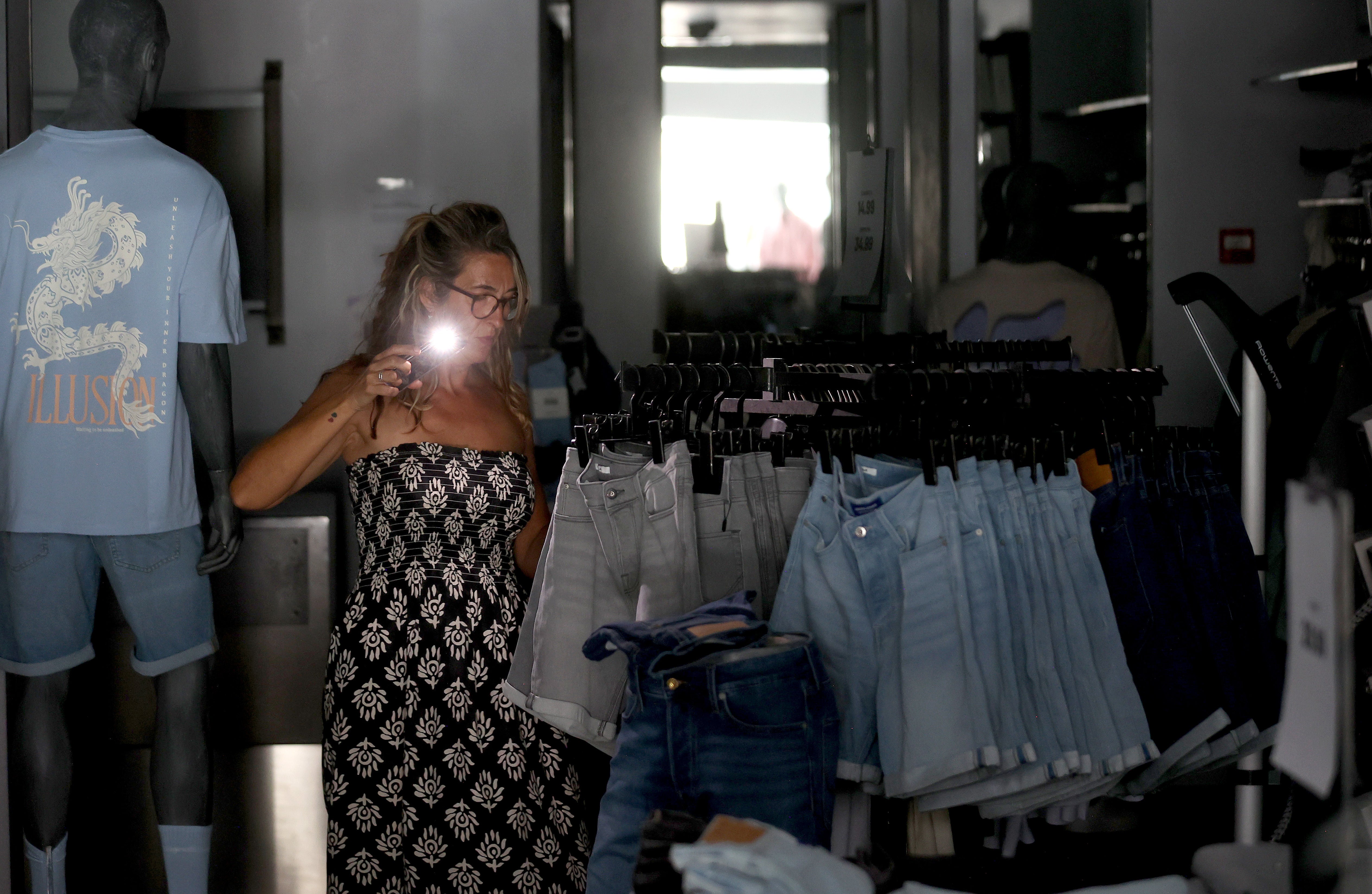 Una mujer alumbra con la linterna de su móvil las prendas de una tienda de ropa.