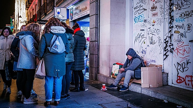 A group of people queue in front of a lottery administration next to a homeless man