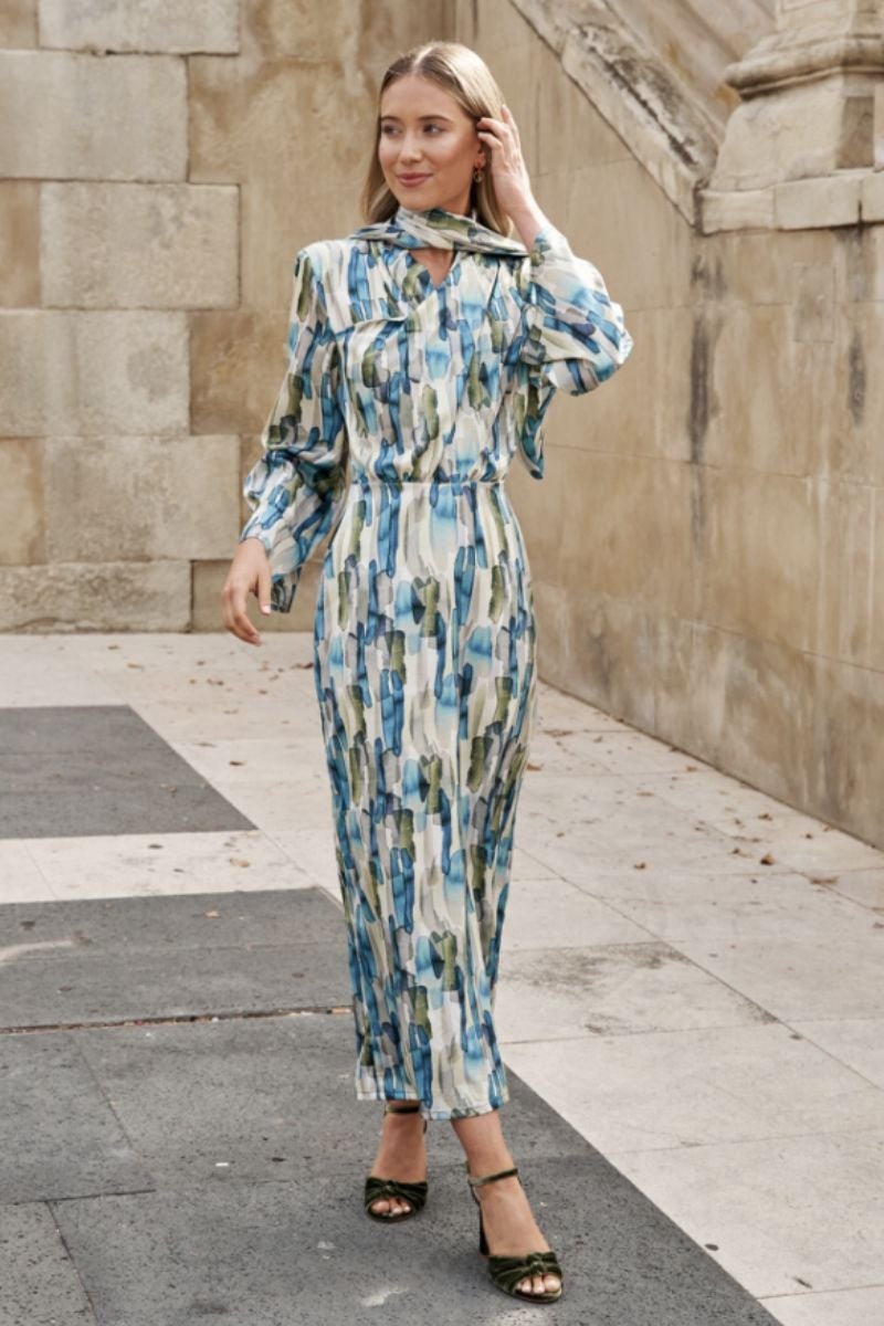 Vestido midi de Cho Atelier con acabado satinado y estampado arty en tonos azules y verdes, y abertura trasera en la falda. Una pieza que cae suavemente sobre la silueta que la equilibra gracias a sus hombreras marcadas y la cintura, que se ajusta suavemente. La prenda incluye un fular al cuello que le aporta un extra de sofisticación. Precio: 151 €.