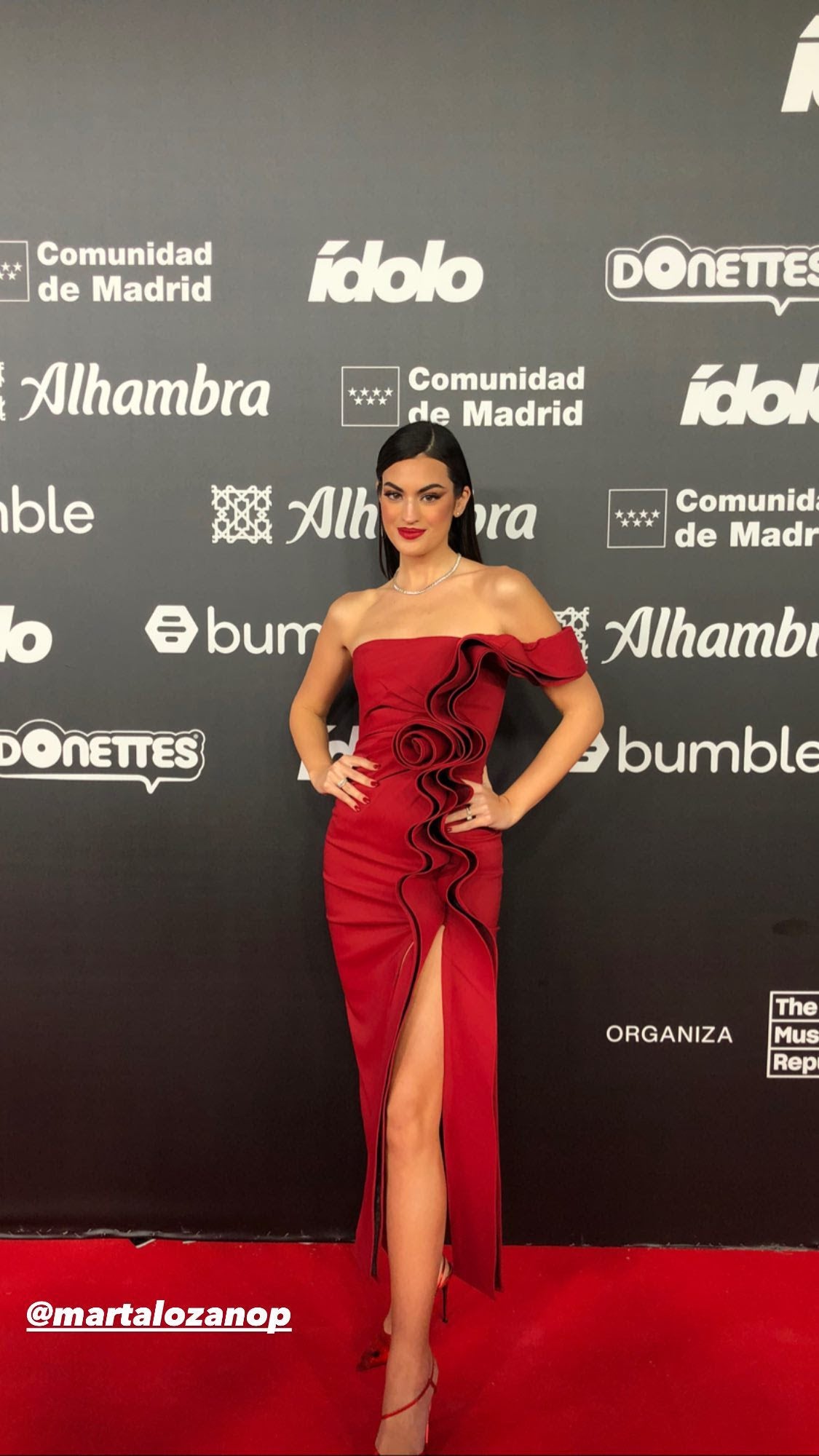 Marta Lozano con vestido midi en color rojo, de escote palabra de honor y con una gran flor de tejido central y volante liso enroscado, de Juan Vidal. Lo combinó con unos zapatos, también de Juan Vidal, en vinilo rojo con strass; y unos pendientes y un collar de la firma Customima.