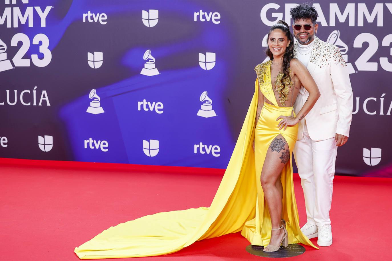 AleMor y Wizzmer con dos estilismos bastante peculiares. Ella ha elegido un escotado vestido amarillo con capa y el un traje blanco con detalles metalizados. 