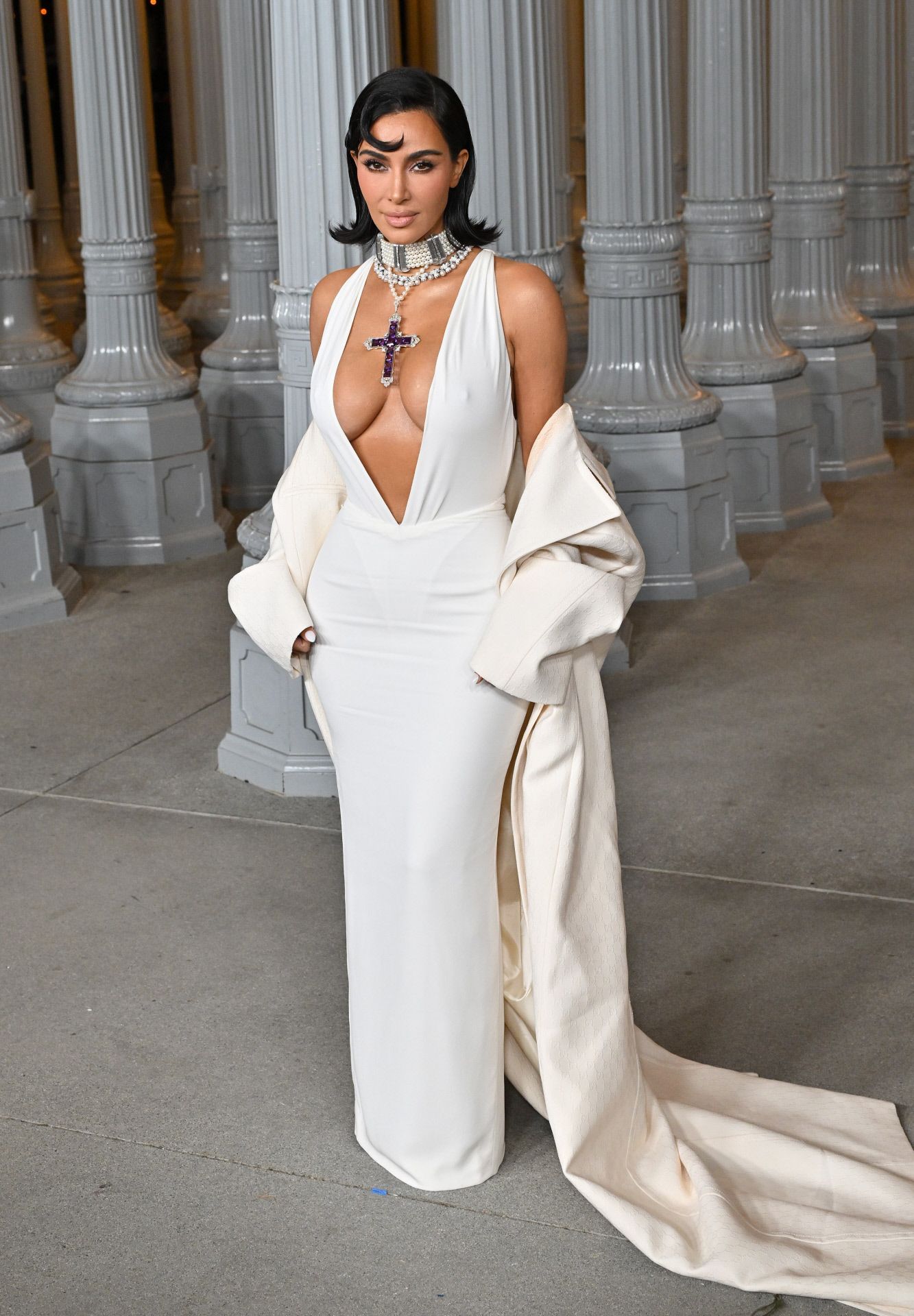 Kim Kardashian lució un vestido blanco con un escote de vértigo, combinado con capa XXL de Gucci.  La empresaria, que lució un bob con las puntas hacia afuera, llevó, además, un collar acompañado de la Cruz de Attallah que lució la princesa de Gales en diversas ocasiones, y que adquirió a través de una subasta de Sotheby's en 2023. 