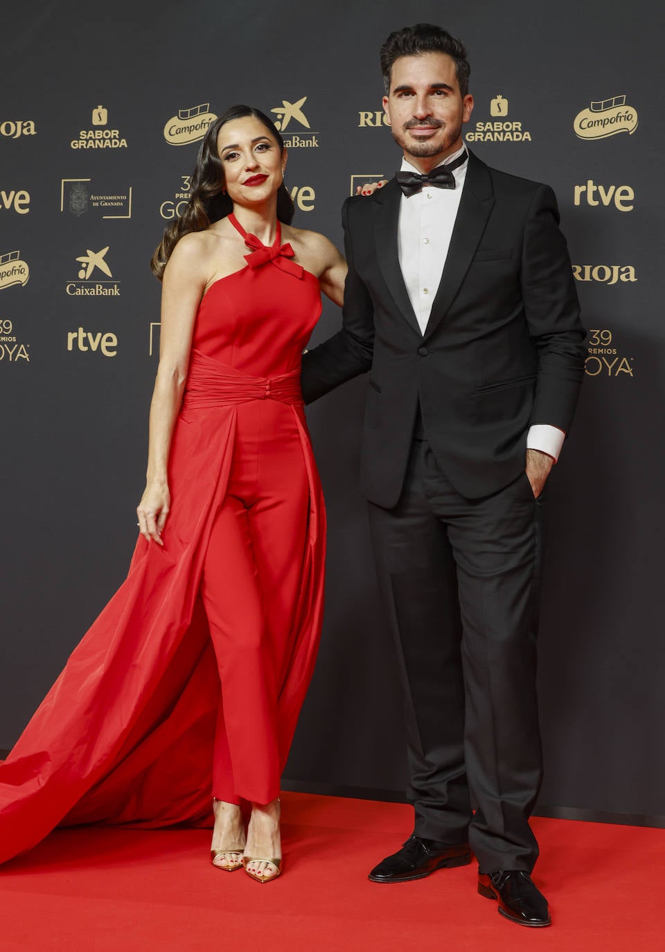 El escritor Javier Castillo , con esmoquin de Armani, zapatos Martinelli y gemelos dorados de Rabat, y su pareja, la influencer Verónica Díaz, con mono largo rojo con escote halter, decorado con un pequeño lazo y espalda descubierta en crepe y satén, combinado con una sobrefalda ajustada a la cintura y con una gran cola de Silvia Fernández. Como complementos joyas de Rabat y sandalias doradas de Giuseppe Zanotti. 
