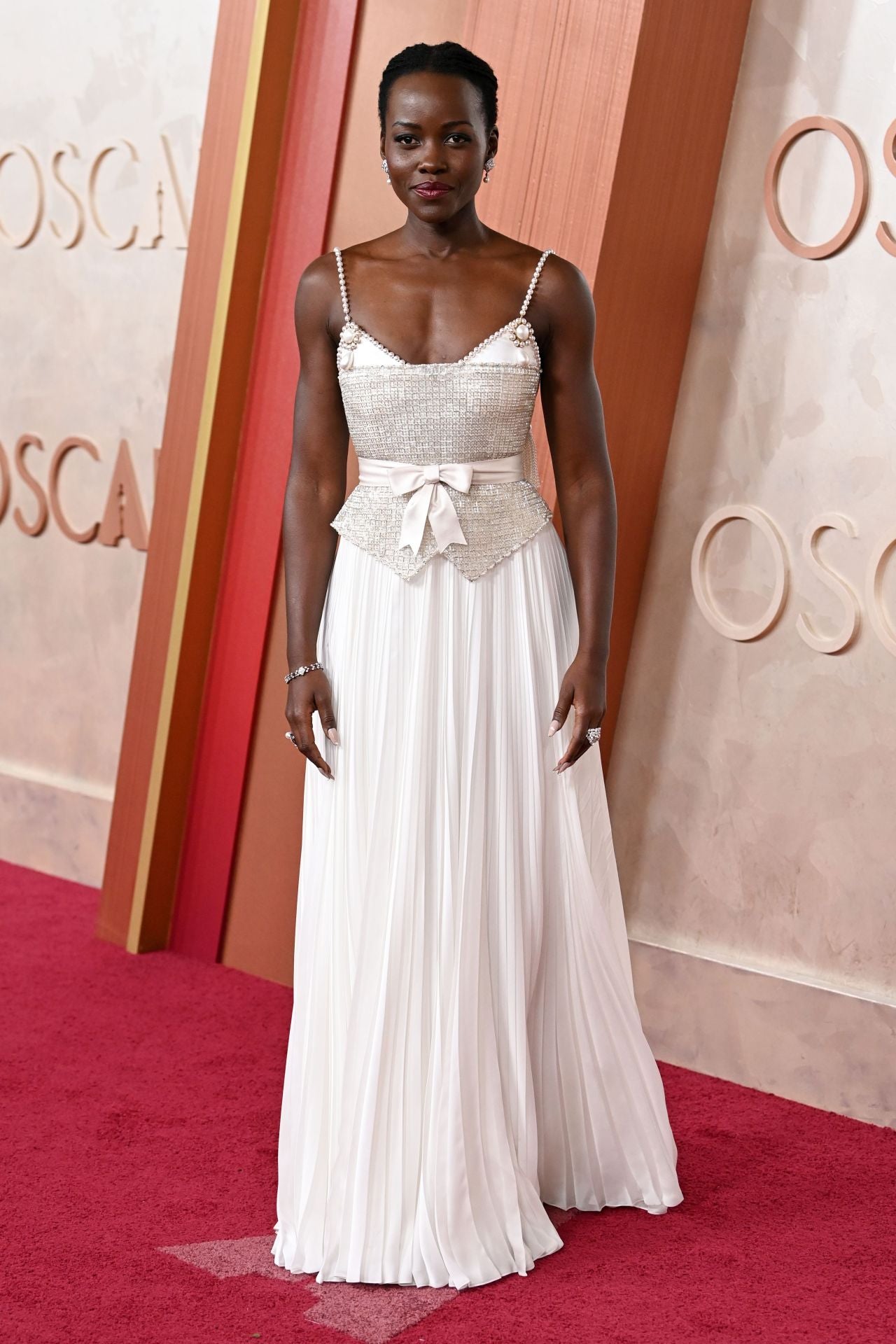 Lupita Nyong'o eligió un vestido blanco de falda plisada con un lazo en la cintura con detalles brillantes y tirantes de perlas de Chanel.