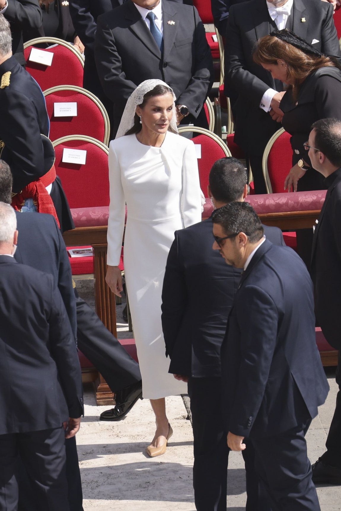 Doña Letizia, que usó el 'privilegio del blanco', estrenó un vestido midi con escote asimétrico creado con drapeado en forma de nudo y confeccionado en crepe de Redondo Brand. Lo combinó con mantilla y bolso de mano, en blanco, y salones bajos en nude de Magrit.