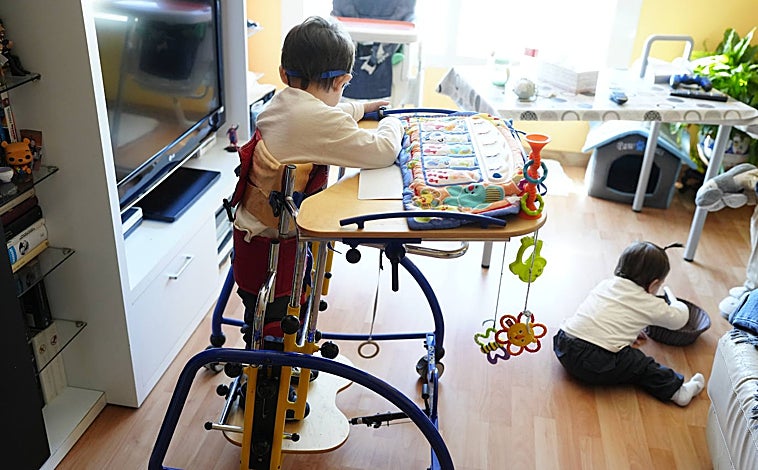 Imagen principal - Eduardo en su bipedestador junto a su hermana Paula (arriba). El pequeño juega en su mesa especial (abajo a la izquierda). María sostiene a su hijo en pie mientras Estefanía juega con él (abajo a la izquierda)