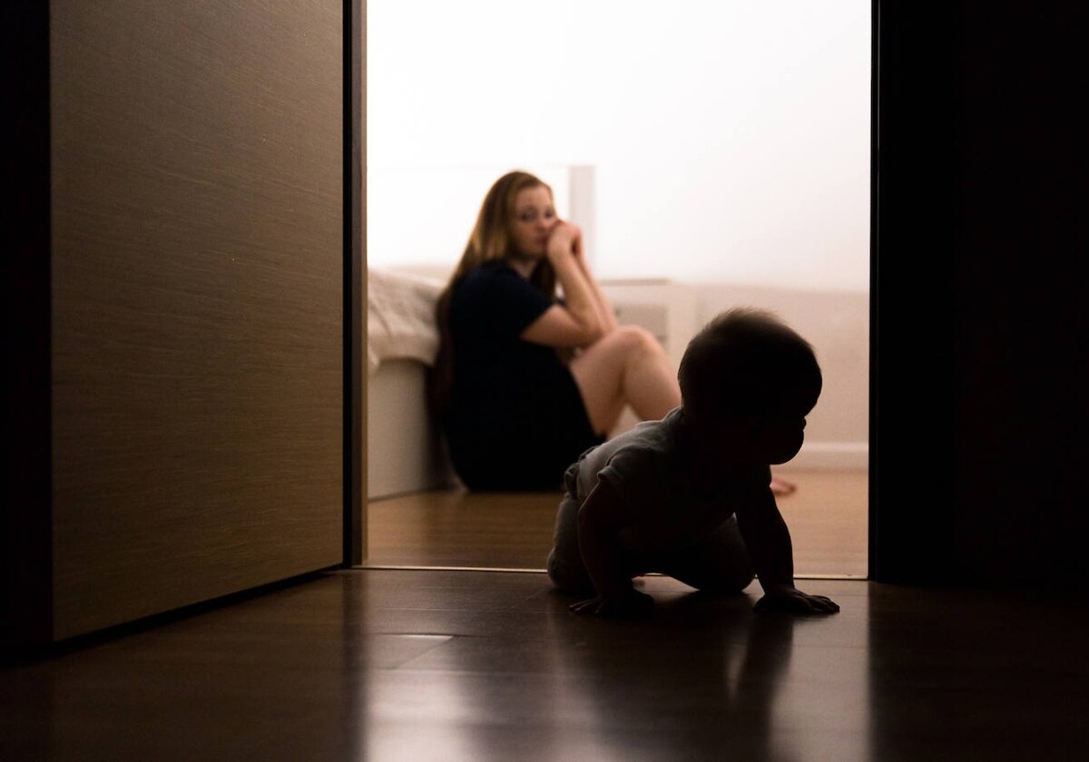 Un bebé gatea mientras su madre de fondo le observa