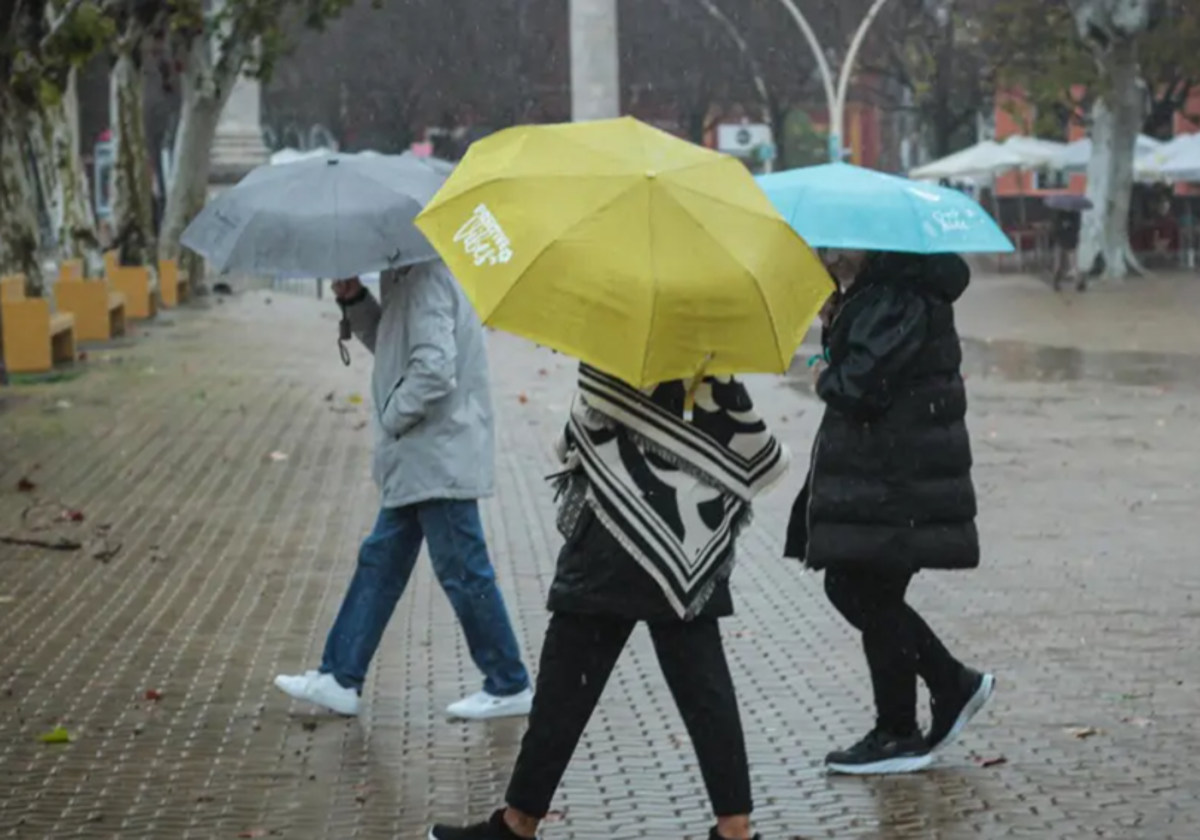 Tres personas, a su paso por la Alameda de Hércules, se protegen de la lluvia con los paraguas en un día de tormenta en Sevilla