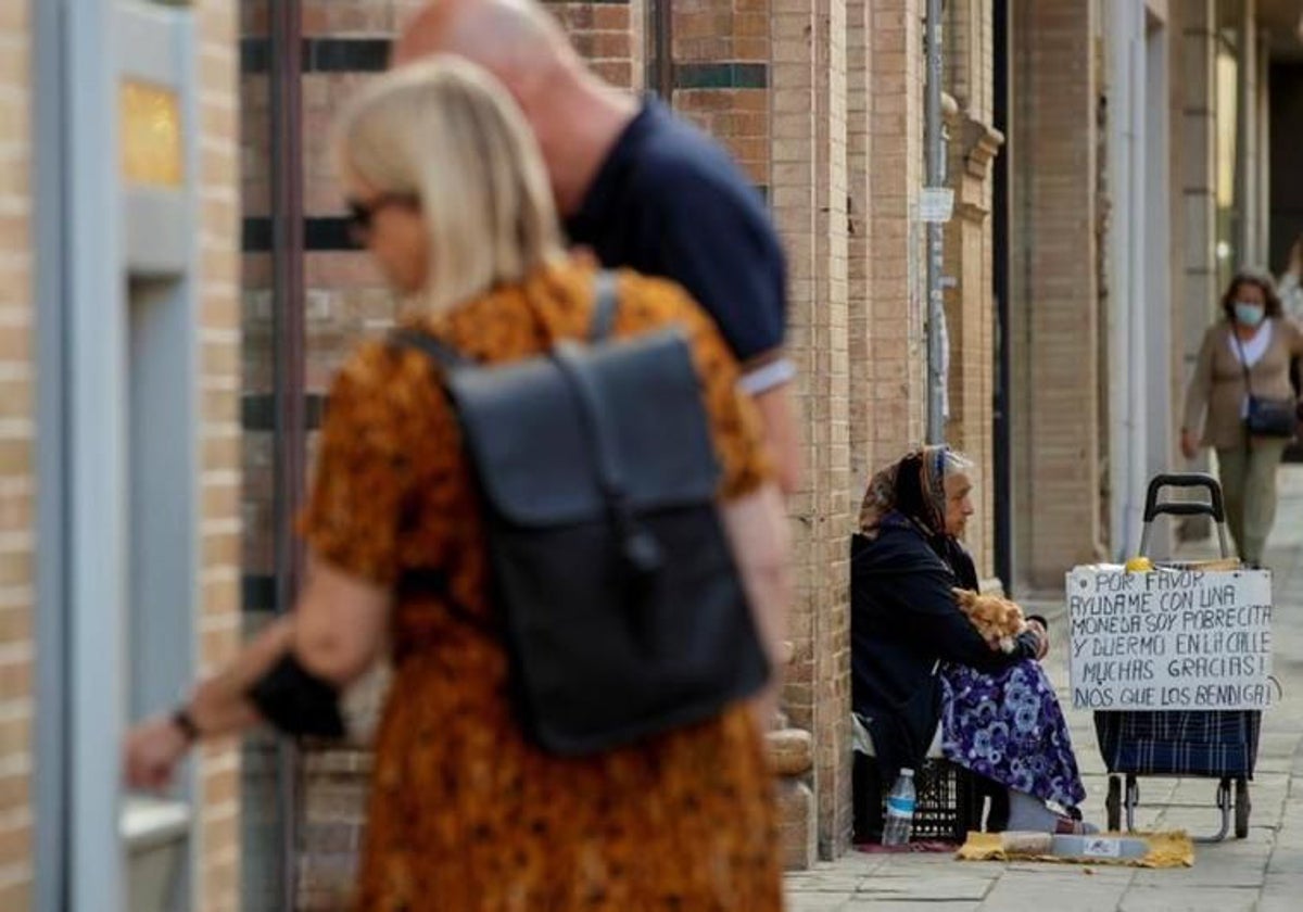 Una mujer pide en la calle, junto a dos turistas que sacan dinero en un cajero