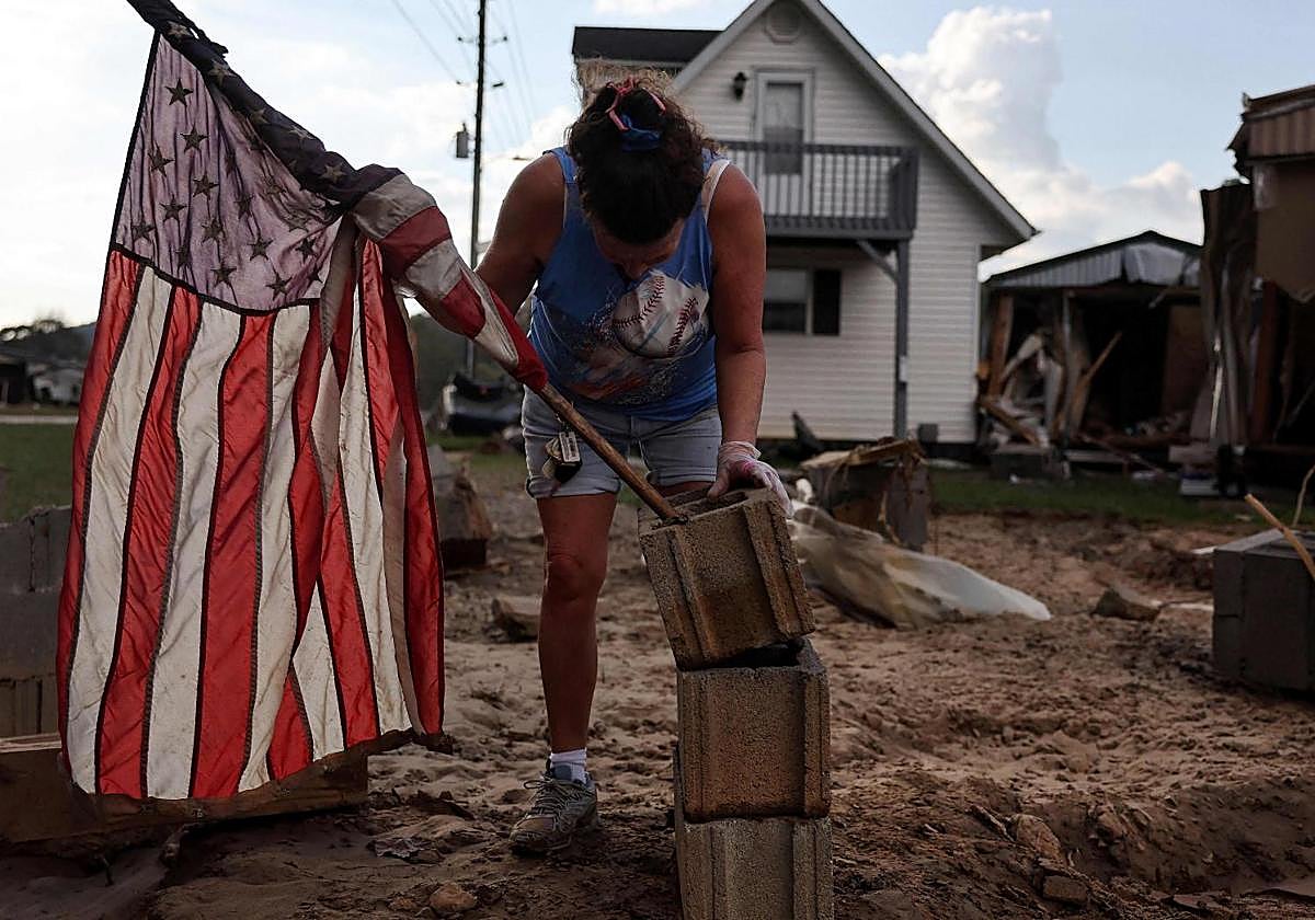Una mujer montando una bandera estadounidense en una pila de bloques de hormigón frente a una casa móvil destruida