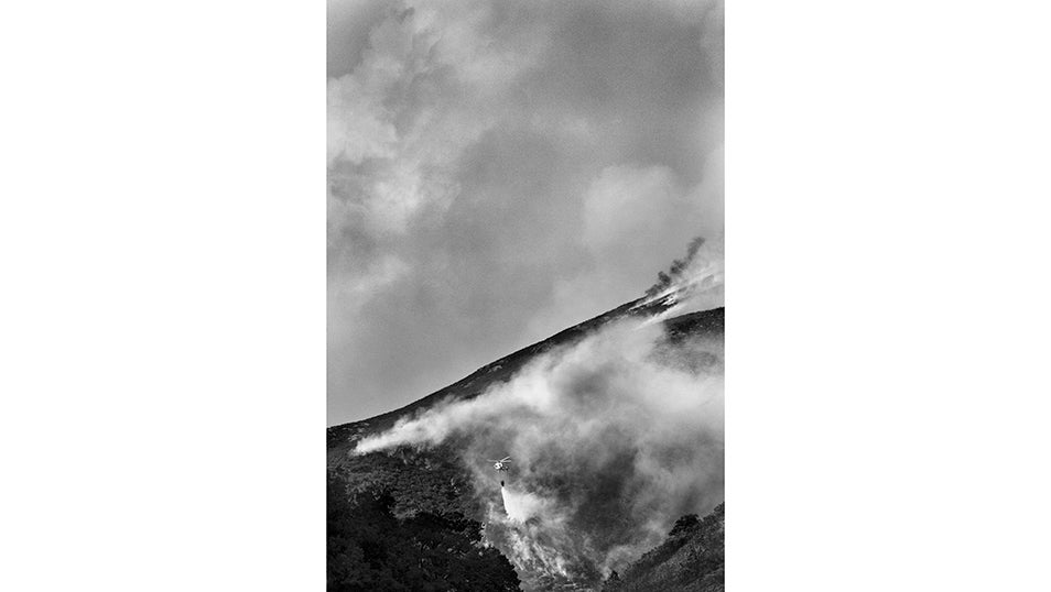 La imagen muestra la ladera de una montaña envuelta en humo y fuego, donde un helicóptero de extinción descarga agua sobre un frente que avanza sin control. El contraste entre la niebla oscura y la silueta de la aeronave refleja la magnitud del incendio y la fragilidad de quienes, desde el aire, intentan frenar el avance de las llamas en un terreno abrupto y hostil. En el valle de Fornela, vecinos y equipos de emergencia han vivido días de incertidumbre y miedo. La orografía de la zona dificulta las maniobras aéreas y convierte cada descarga en un esfuerzo titánico para contener un fuego que amenaza pueblos enteros como San Pedro de Paradela, Cariseda y Peranzanes. La coordinación entre brigadistas, bomberos y voluntarios se complementa con los medios aéreos, que tratan de enfriar las zonas más expuestas para dar un respiro a quienes combaten en tierra. El incendio, declarado de nivel 2 por su peligrosidad, se originó en Anllares del Sil y se extendió rápidamente hasta alcanzar el valle. Durante más de diez días ha puesto en jaque a toda la comarca del Bierzo y ha obligado a evacuar a cientos de personas. En estas montañas, donde generaciones han trabajado la tierra y levantado sus casas piedra a piedra, la lucha contra el fuego no solo busca salvar construcciones: se trata de defender la memoria, el esfuerzo y el futuro de quienes llaman hogar al valle de Fornela.