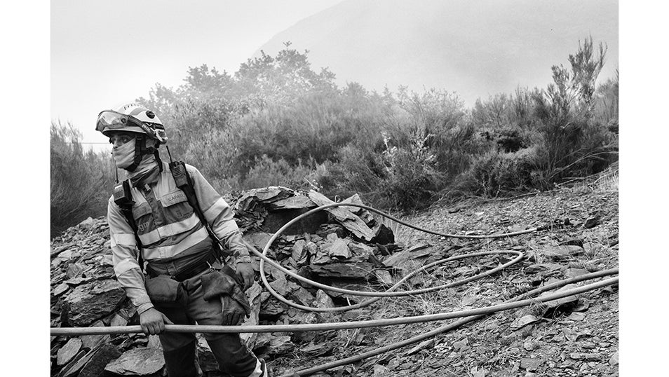 Un bombero sostiene una manguera mientras observa el avance del fuego en la ladera. El humo cubre parte del horizonte y convierte cada movimiento en una tarea lenta y vigilante. La tensión se refleja en la postura: preparado para actuar en cualquier momento. En el valle de Fornela, los equipos trabajaron durante horas en condiciones extremas para contener las llamas y evitar que alcanzaran los pueblos cercanos. La presión constante por mantener la línea de defensa fue clave para impedir que el incendio cruzara la carretera y arrasara todo el valle.