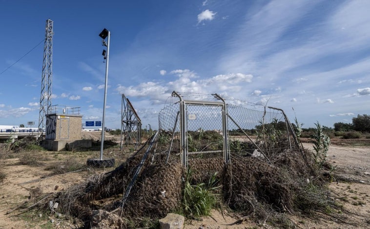 Imagen principal - En la imagen superior, la zona de la valla de protección del caudalímetro que no fue derribada por la riada todavía tiene cañas y matorrales acumulados, el resto está en el suelo, semienterrada. En la segunda imagen se puede observar la caseta del caudalímetro, a la izquierda, y el puente de la A3 que pasa por encima del barranco del Poyo. Un cable une el poste de la instalación con el sensor láser  instalado en el puente. En la tercera imagen, un primer plano de la caseta y las placas solares que la alimentan,