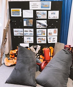 Imagem secundária 2: A cena principal é a filmagem do resgate do último corpo encontrado de um dos filhos mais novos da família na chegada a Labuan Bajo, na Indonésia. Abaixo segue uma folha com informações sobre a tripulação e passageiros do KM Putri Sakina, com os nomes dos menores riscados e a presença do capitão Lukman, que, segundo os investigadores, nunca esteve no navio; Ao lado está uma imagem dos restos resgatados do navio e um quadro com informações nas quais a polícia indonésia está trabalhando.