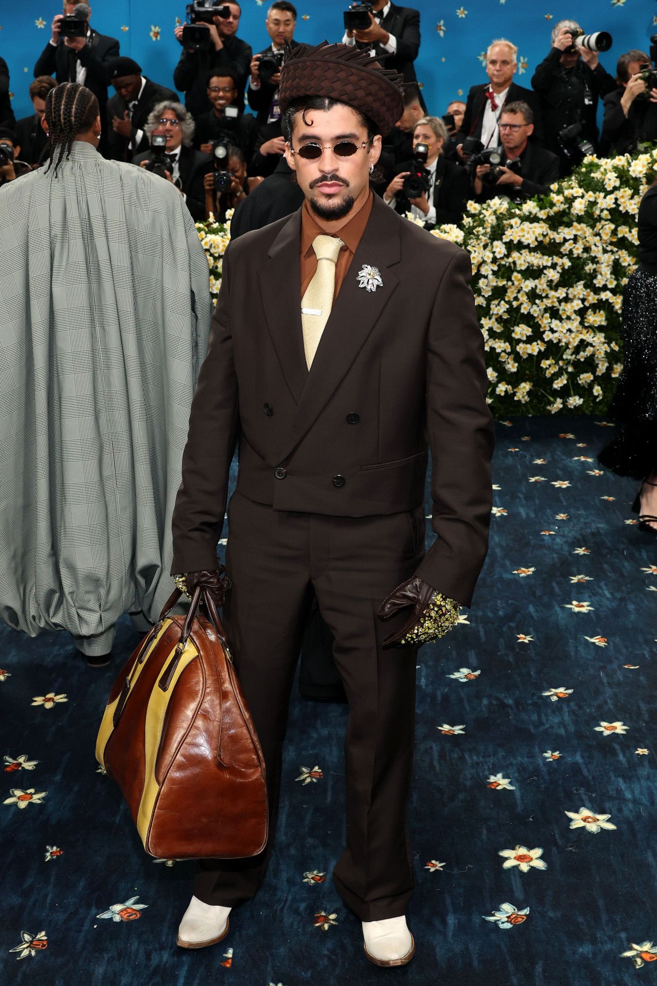 Bud Bunny llevó un traje de color marrón chocolate, formado por chaqueta corta cruzada y pantalón recto, que combinó con camisa y corbata en crema, a juego con los zapatos. El cantante añadió diversos complementos a su look: un broche en la solapa de Cartier, guantes de piel con incrustaciones metálicas, un original sombrero hecho a mano en Puerto Rico y un bolso XXL de Prada. 