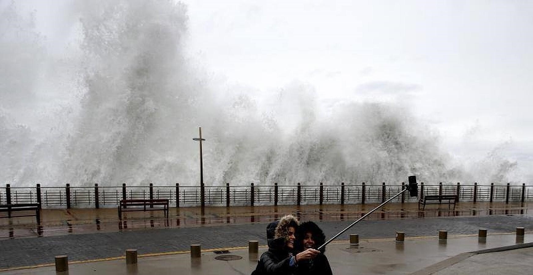 Dos jóvenes se fotografían con una ola rompiendo en el Paseo Nuevo de San Sebastián