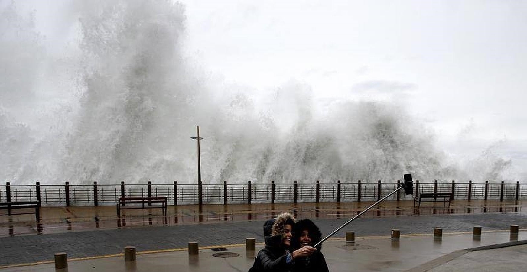 Dos jóvenes se fotografían con una ola rompiendo en el Paseo Nuevo de San Sebastián. 