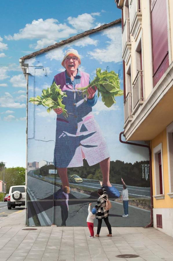 Lola, la greleira de 50 pies. «Se le llama la ruta del grelo a un tramo de carretera antes de entrar en Ordes (La Coruña). Las mujeres venden sus grelos en pequeños puestos cerca de la carretera o en la puerta de sus casas. Lola, que así se llama la modelo, es la abuela de una amiga de Cambre. Sujetando un manojo de grelos en cada mano, hice la versión gallega de «El ataque de la mujer de 50 pies», una película de serie B de los cincuenta. Los grelos de ordes son mucho más potentes que la espinacas de Popeye», dice el autor sobre este mural.