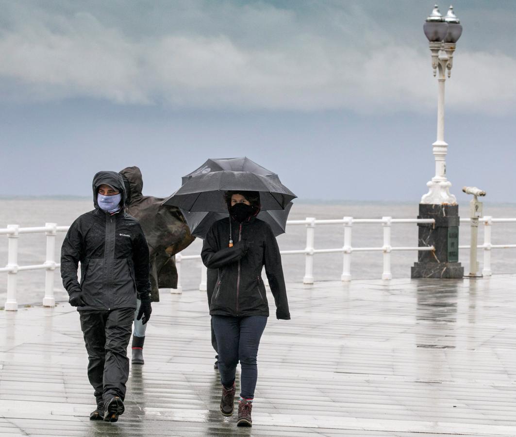 Un grupo de personas paseando por la playa de San Lorenzo de Gijon, bajo un fuerte chubasco y viento. 