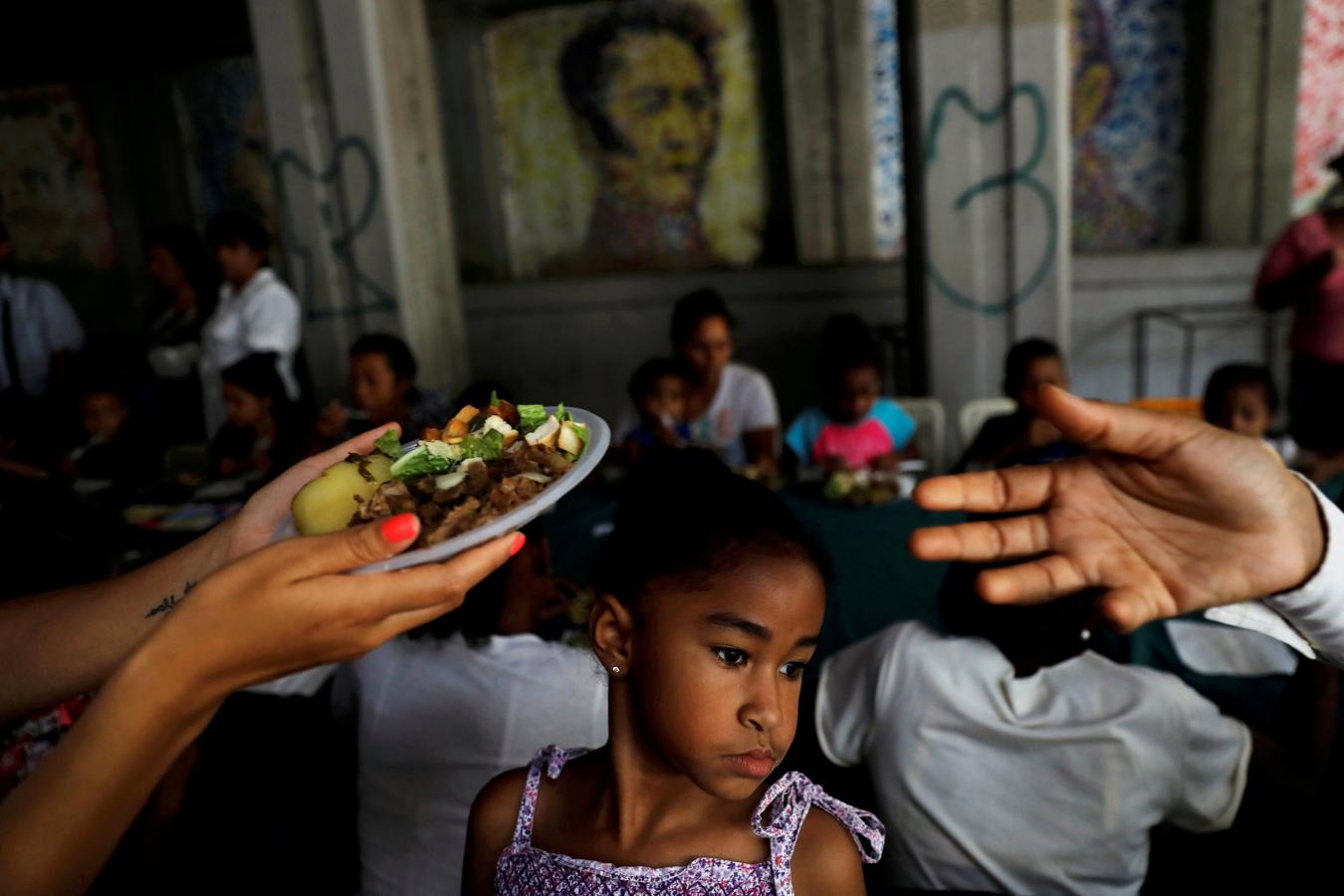 Un niño observa como voluntarios del candidato presidencial venezolano Javier Bertucci del partido «Esperanza por el Cambio» entregan platos de comida a mujeres y niños como parte de la celebración del Día de la Madre, durante un mitin de campaña en Caracas, Venezuela, el 13 de mayo. 2018.. 