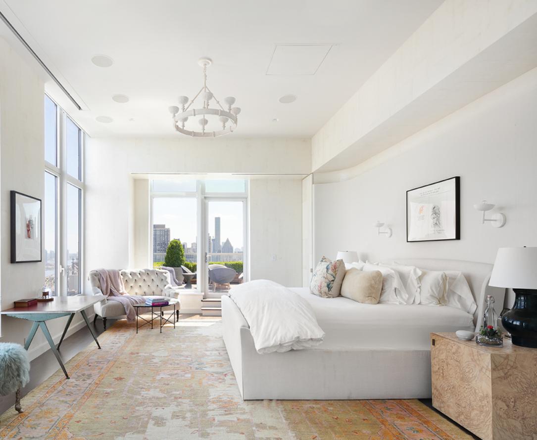 El espectacular ático de Jennifer Lawrence en Nueva York. El dormitorio principal dispone de un gran vestidor y cuarto de baño. Una gran alfombra destaca sobre el resto del decorado y mobiliario que sigue los tonos claros predominantes del resto de la casa. Al fondo, un rincón de lectura con un pequeño sofá con capitoné y salida directa a la terraza.