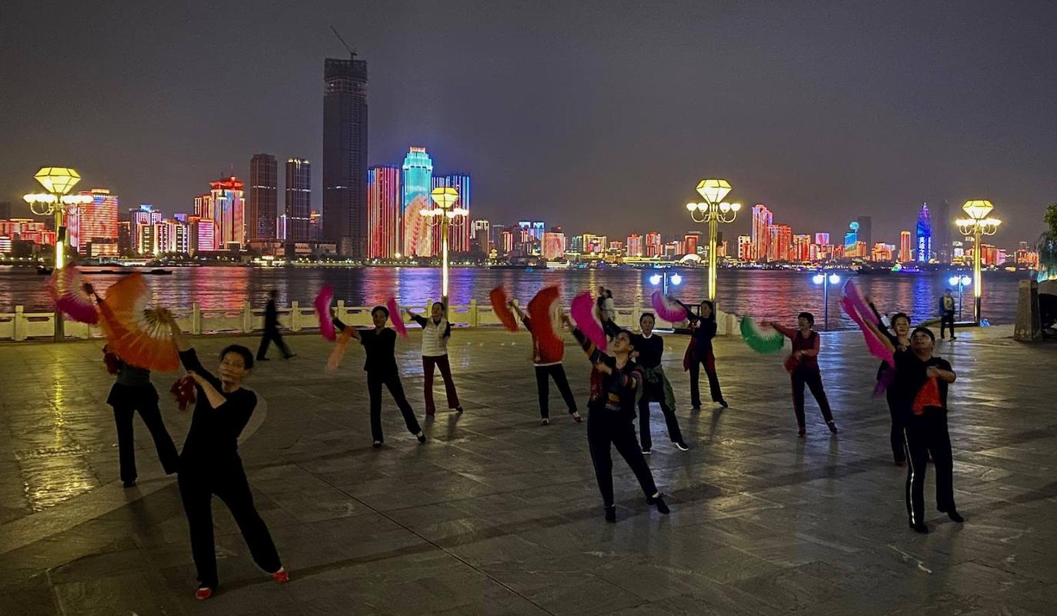 Como es costumbre en China, un grupo de mujeres baila al anochecer en un parque a orillas del Yangtsé, cuyos rascacielos se iluminan de colores para dar ánimos a los habitantes de Wuhan. 