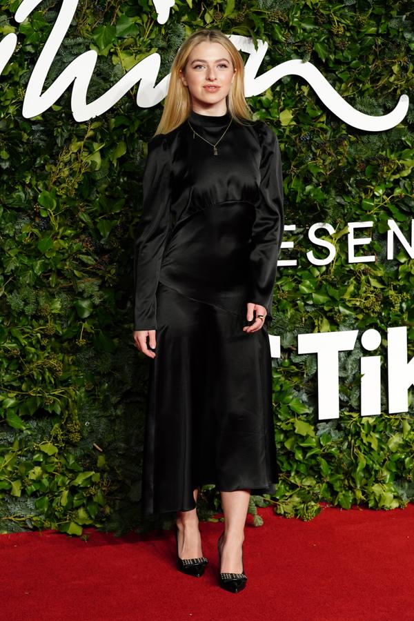 Anais Gallagher - Alfombra roja de los British Fashion Awards. Lució un discreto vestido negro midi confeccionado en satén con cuello cisne y mangas abullonadas cuya falda presentaba una ligera asimetría en el bajo.