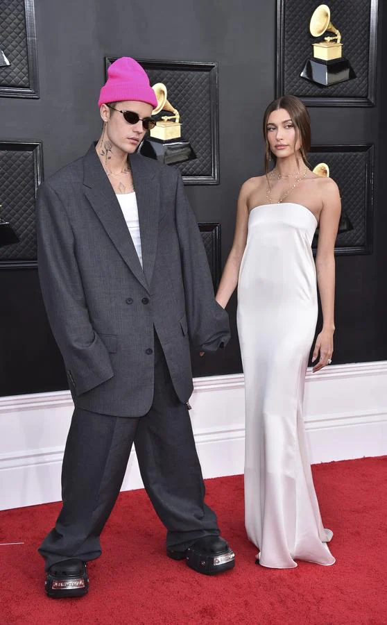 Justin y Hiley Bieber - Alfombra roja de los Grammy 2022. El cantante llevó un traje gris oversize  con una gorra de lana rosa mientras que ella apostó por un minimalista vestido lencero de satén blanco con escote palabra de honor de Saint Laurent.