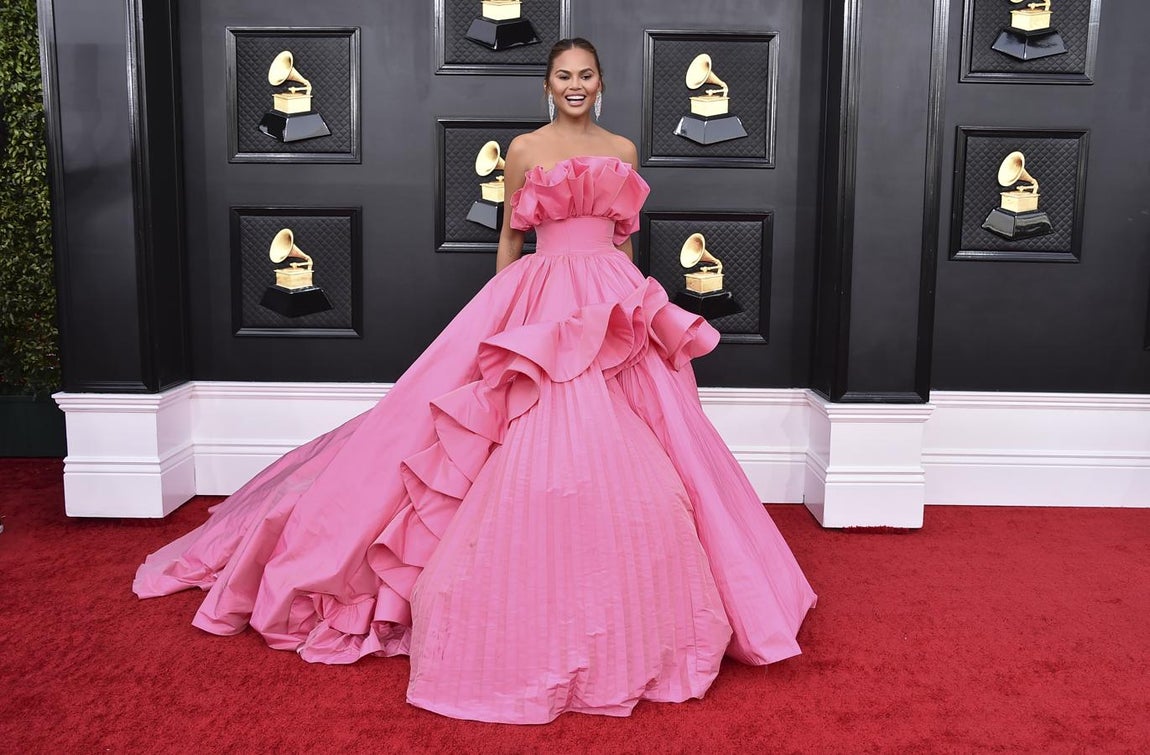 Chrissy Teigen - Alfombra roja de los Grammy 2022. Una de las más llamativas y coloridas de la noche con un voluminoso vestido rosa con volantes XXL y escote palabra de honor con otro gran volante de Nicole + Felicia.