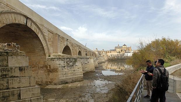 Turistas contemplan el Puente Romano desde una zona próxima a la orilla del río