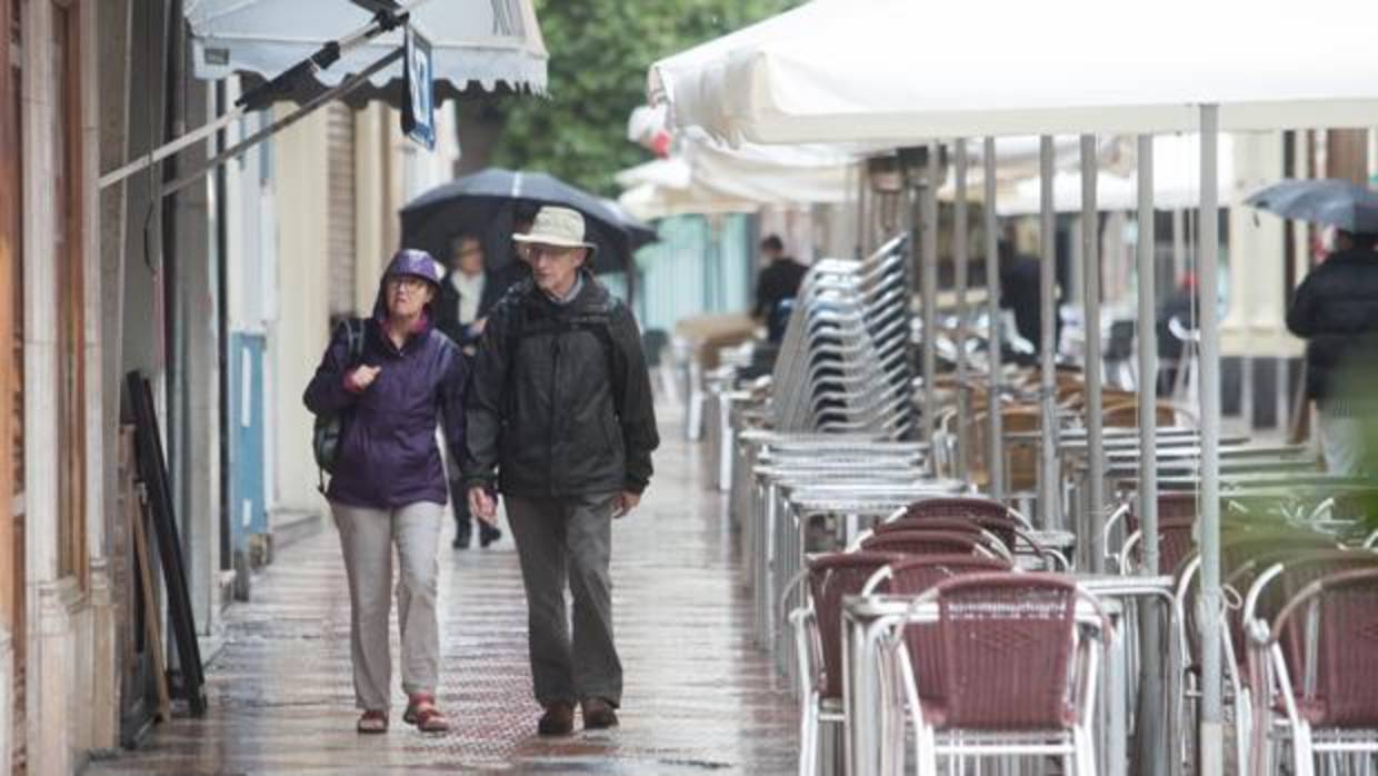 Una pareja de turistas pasean junto a una terraza vacía por la lluvia