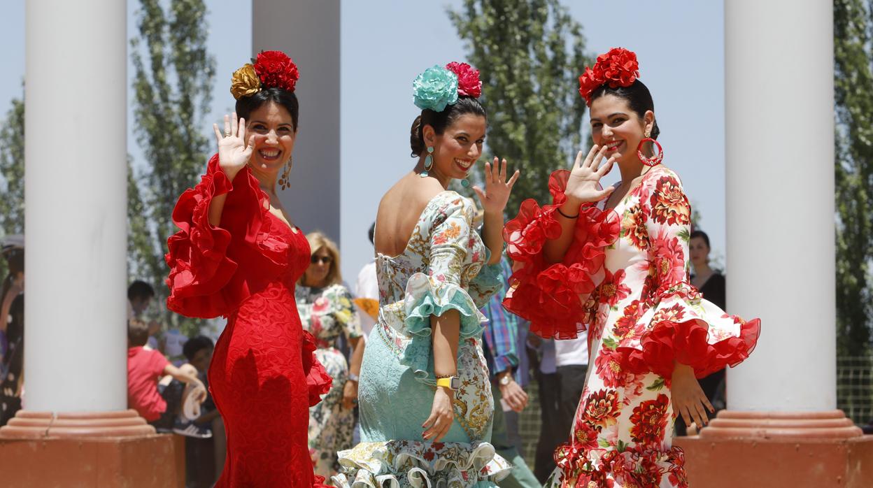 Tres jóvenes vestidas de gitana saludan junto a la portada de la Feria