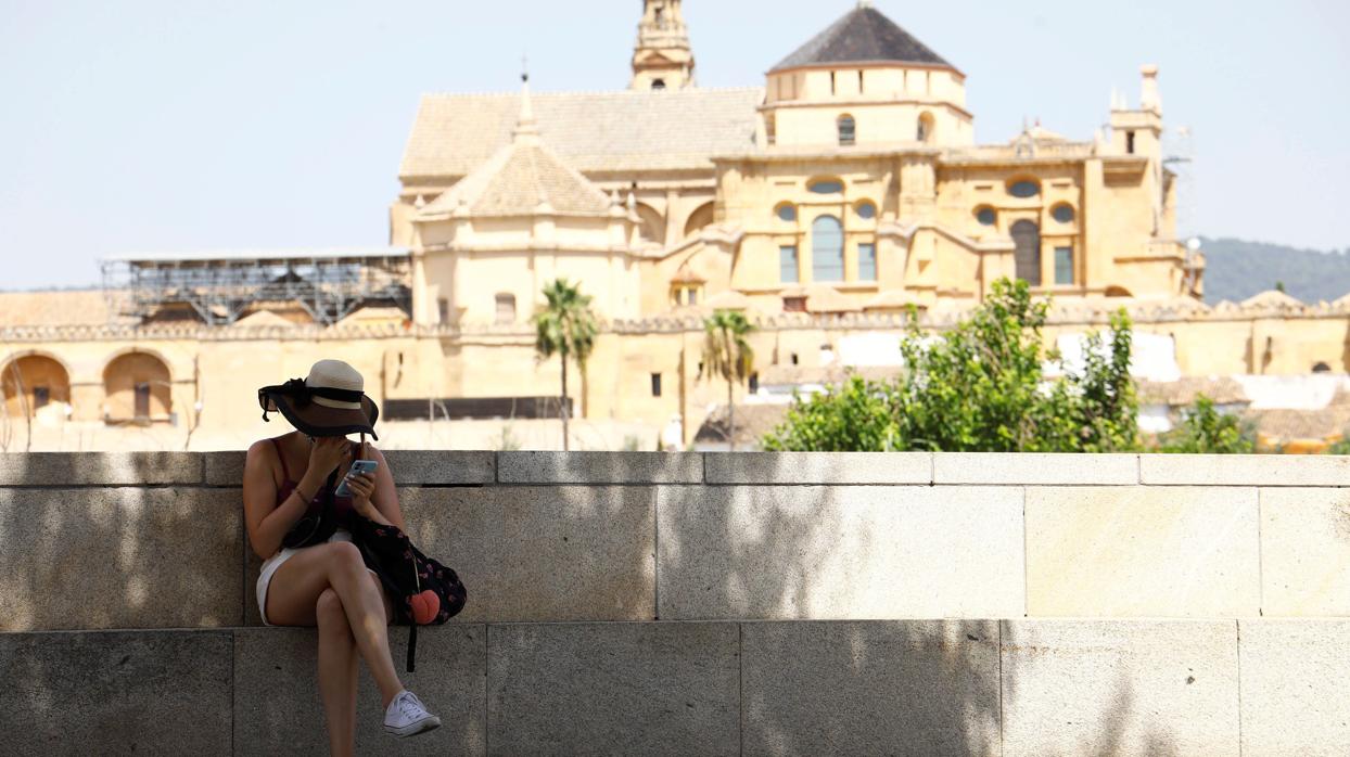 Una turista observa su móvil sentada en la margen izquierda del río Guadalquivir frente a la Mezquita-Catedral