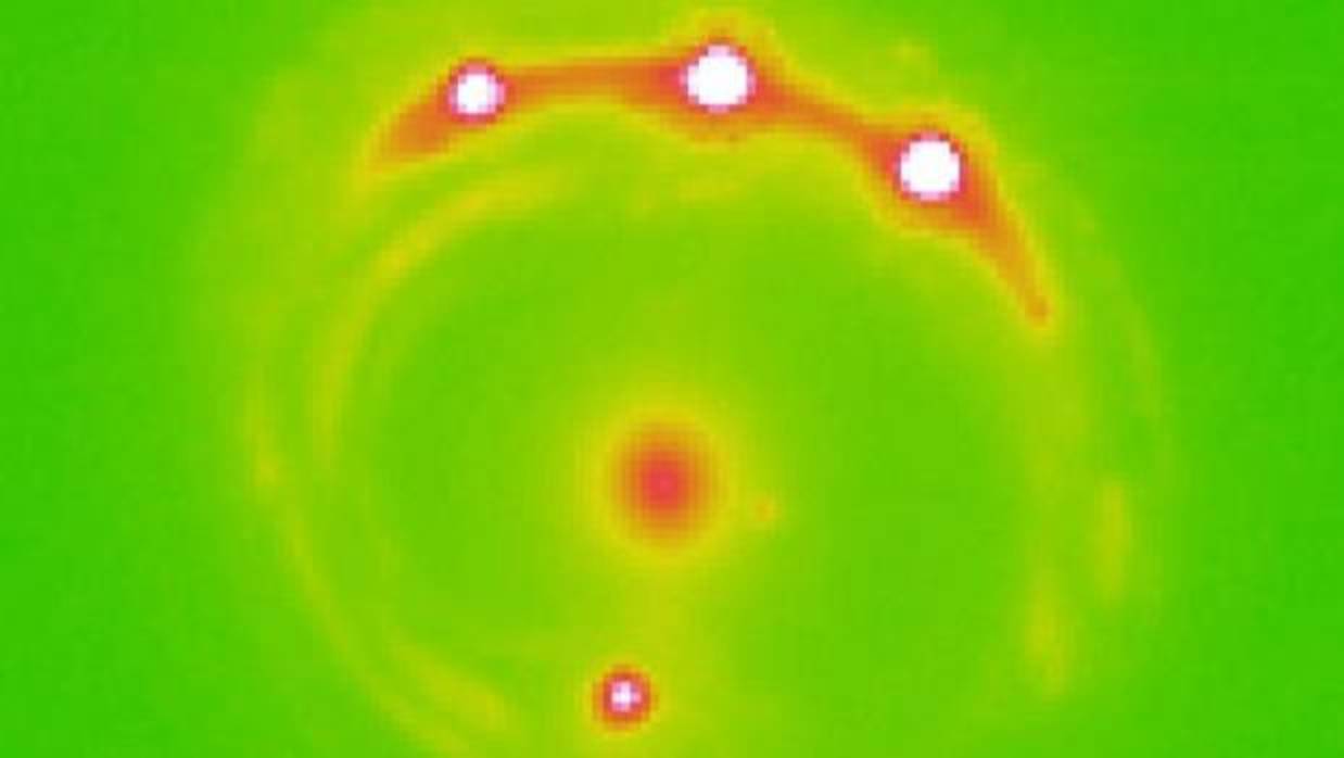Imagen de la lente gravitacional RX J1131-1231. La galaxia que hace de lente está en el centro, desviando la luz de los cuatro cuásares lejanos que aparecen en el borde del disco de luz