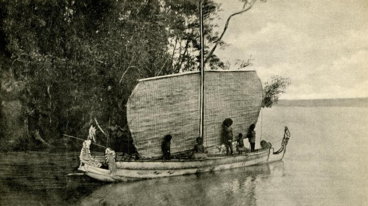 Vista de una gran extensión de agua y una orilla cubierta de árboles, una canoa tradicional con vela está en el agua, varias personas están de pie en la canoa; Isla Woodlark, Papúa Nueva Guinea
