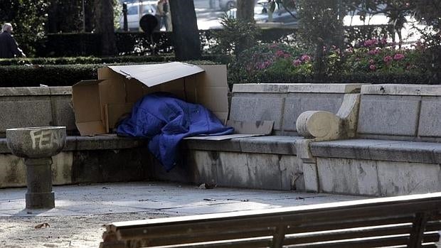 Una persona sin hogar duerme entre cartones en uno de los bancos de piedra del Paseo del Prado