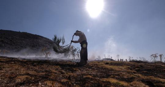 Un vecino de Riofrío golpea la tierra humeante, a pocos metros de los bomberos, que riegan para evitar que se reaviven las brasas