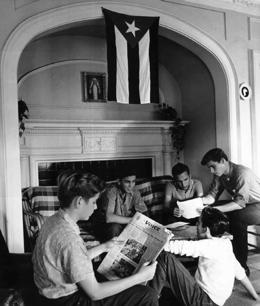 Varios chicos leen la prensa en un salón presidido por la bandera cubana