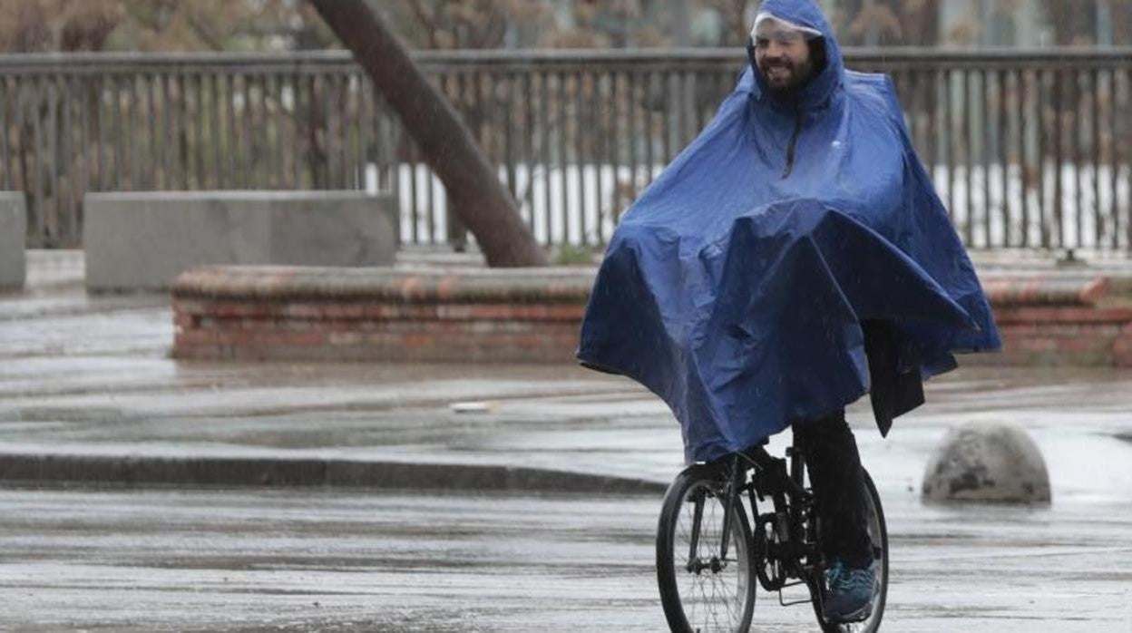 Un hombre pasea en bicicleta por Sevilla protegido de la lluvia con un chubasquero